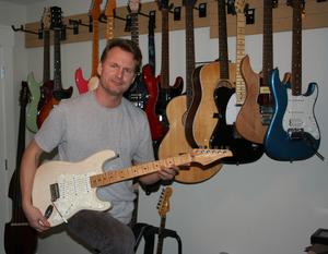 Jim Rhindress holding a cream-coloured electric guitar with many other guitars hanging on the wall behind him